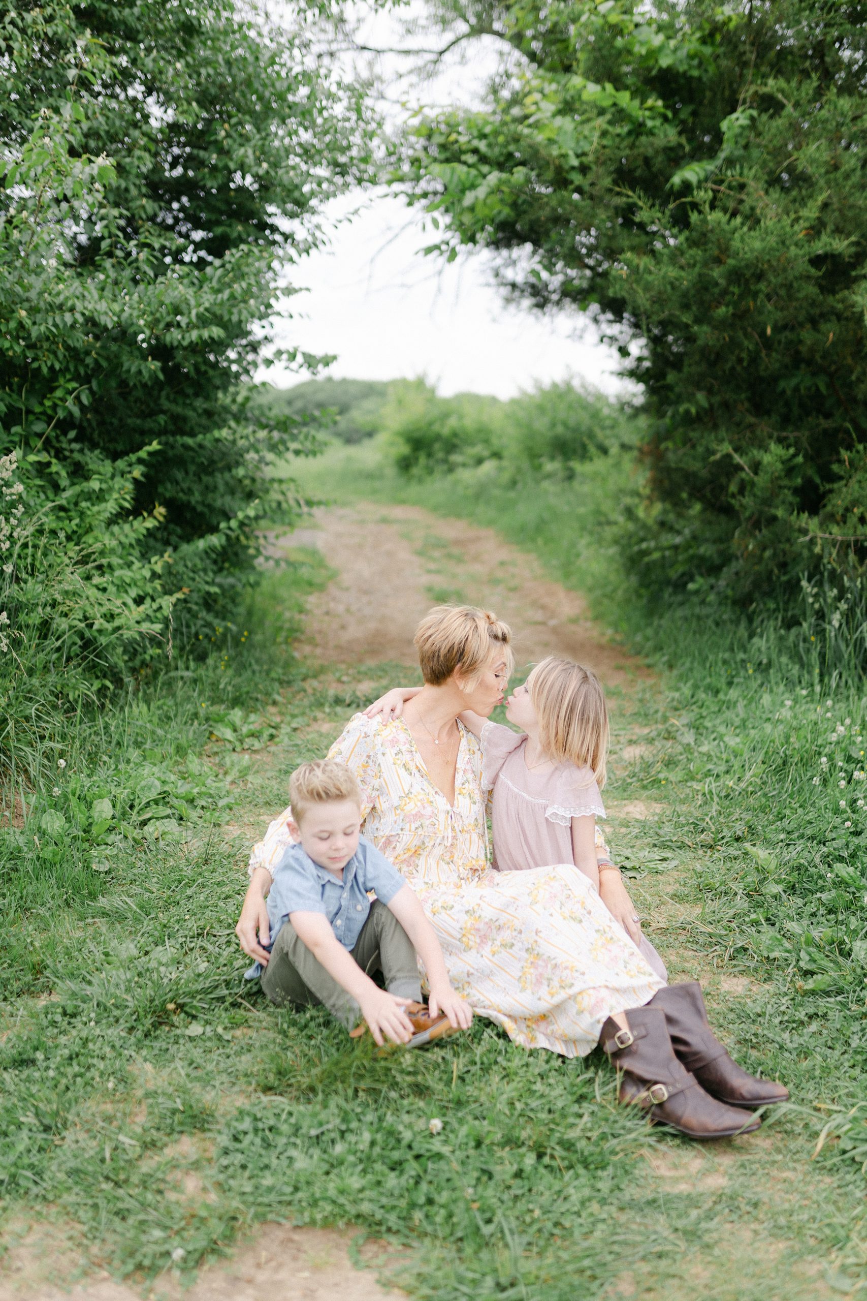 mom sits in grass with two kids and daughter leans up to kiss her mom